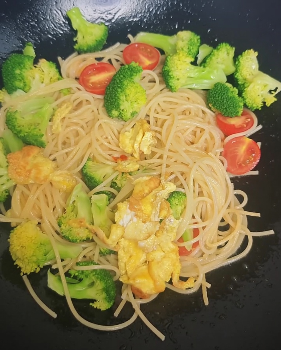 Seasoning being sprinkled from a packet over the spaghetti, broccoli, and cherry tomatoes in a wok.
