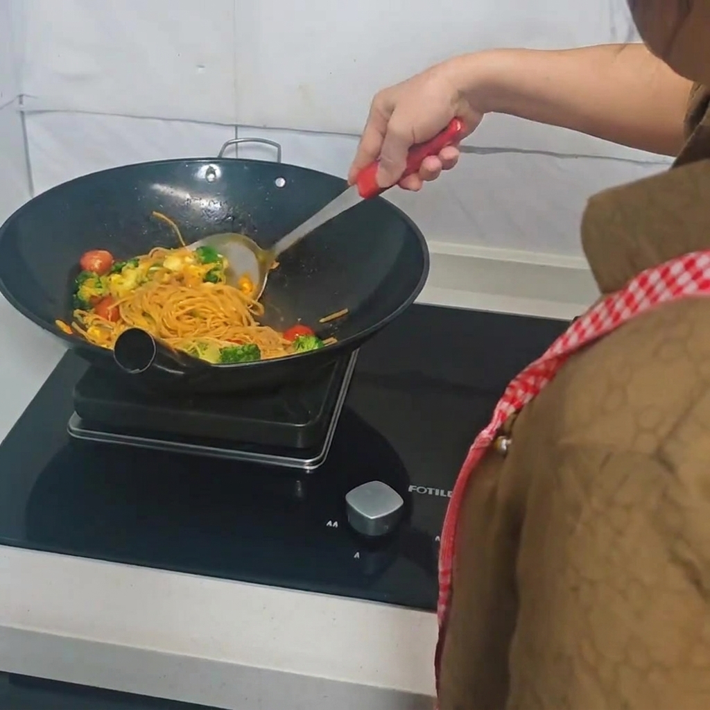 A person using a spatula to stir-fry spaghetti, broccoli, cherry tomatoes, and eggs in a wok.
