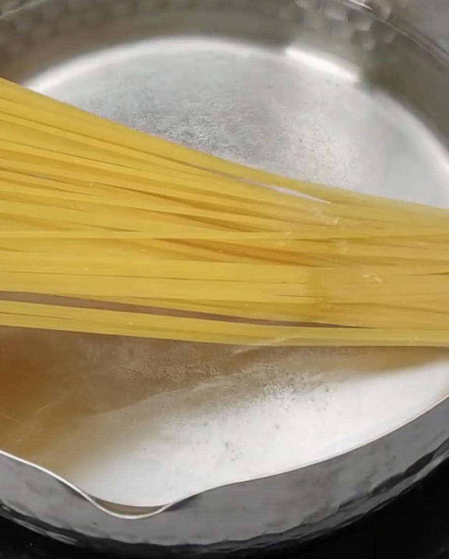 Dry long pasta noodles being lowered into a stainless steel pot filled with vigorously boiling water.