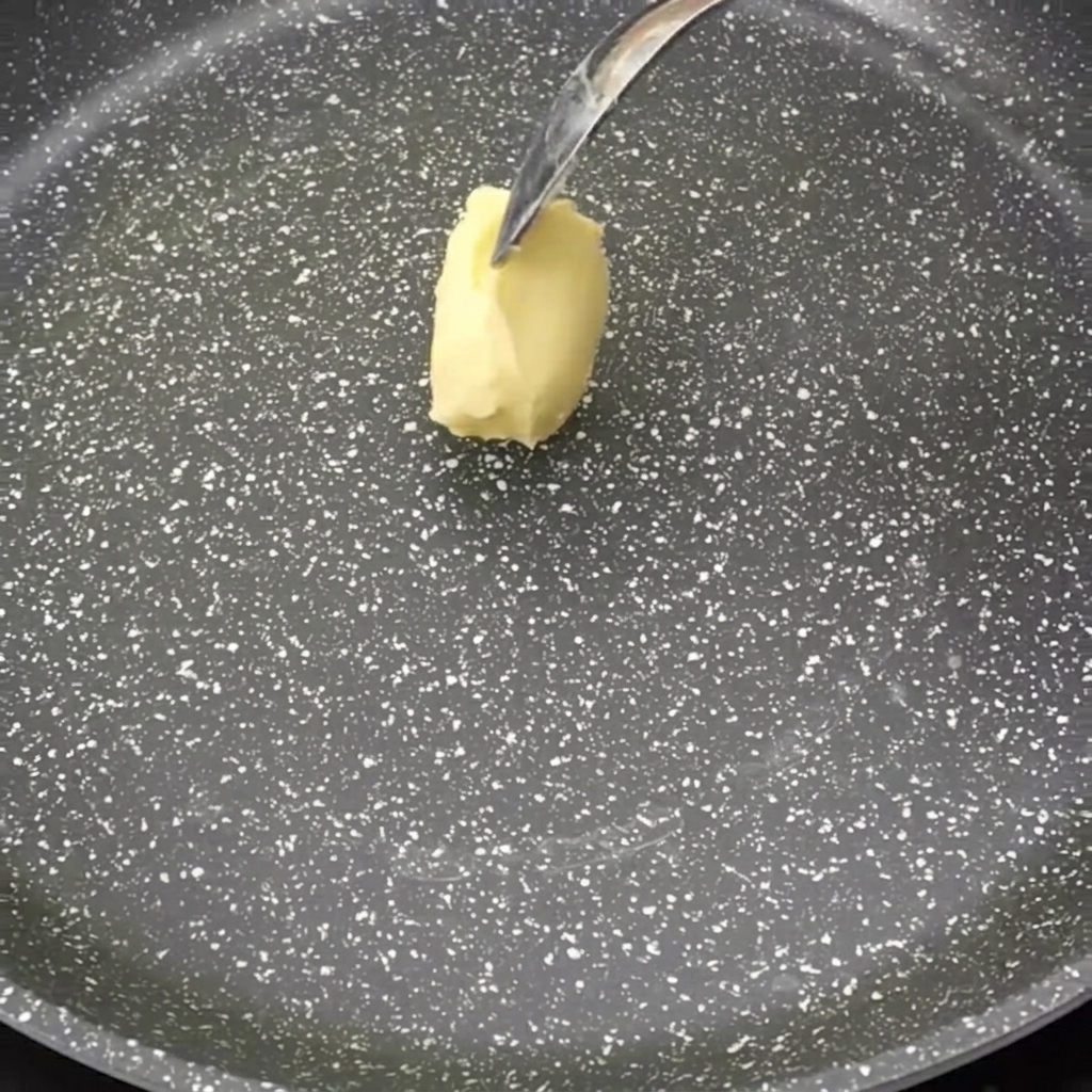 A small pat of pale yellow butter melting off a spoon into a dark speckled non-stick skillet.