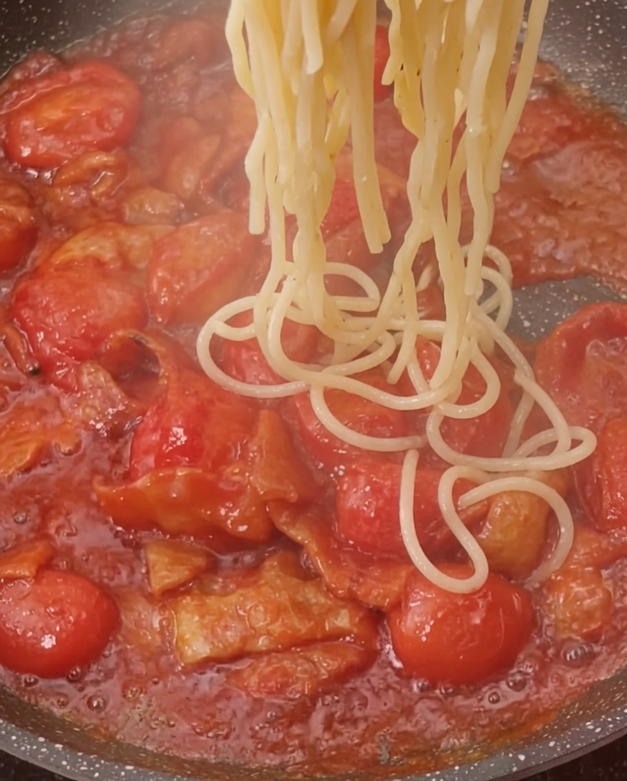 Freshly boiled pasta being added to a pan filled with a simmering tomato and bacon sauce.