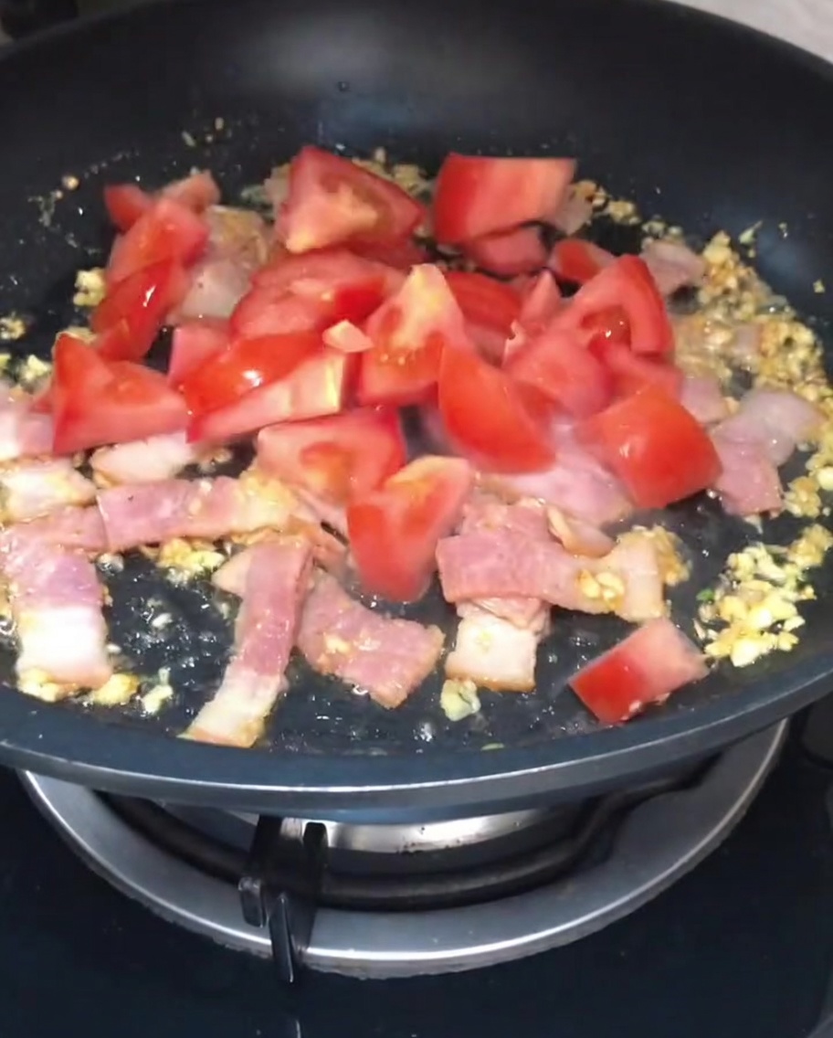 Freshly chopped red tomato chunks being added to the sautéing bacon and garlic in a pan.