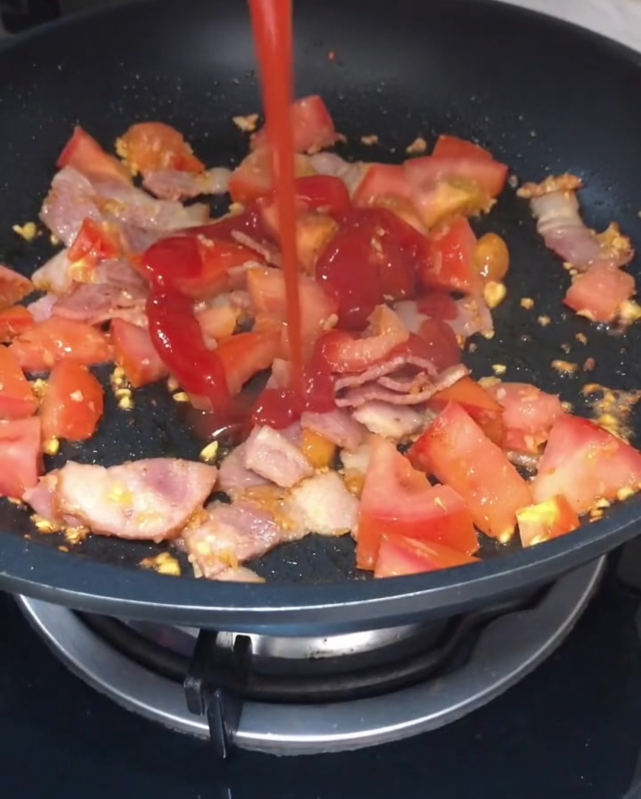 Ketchup and light soy sauce being drizzled over the sautéed tomato and bacon mixture in the pan.