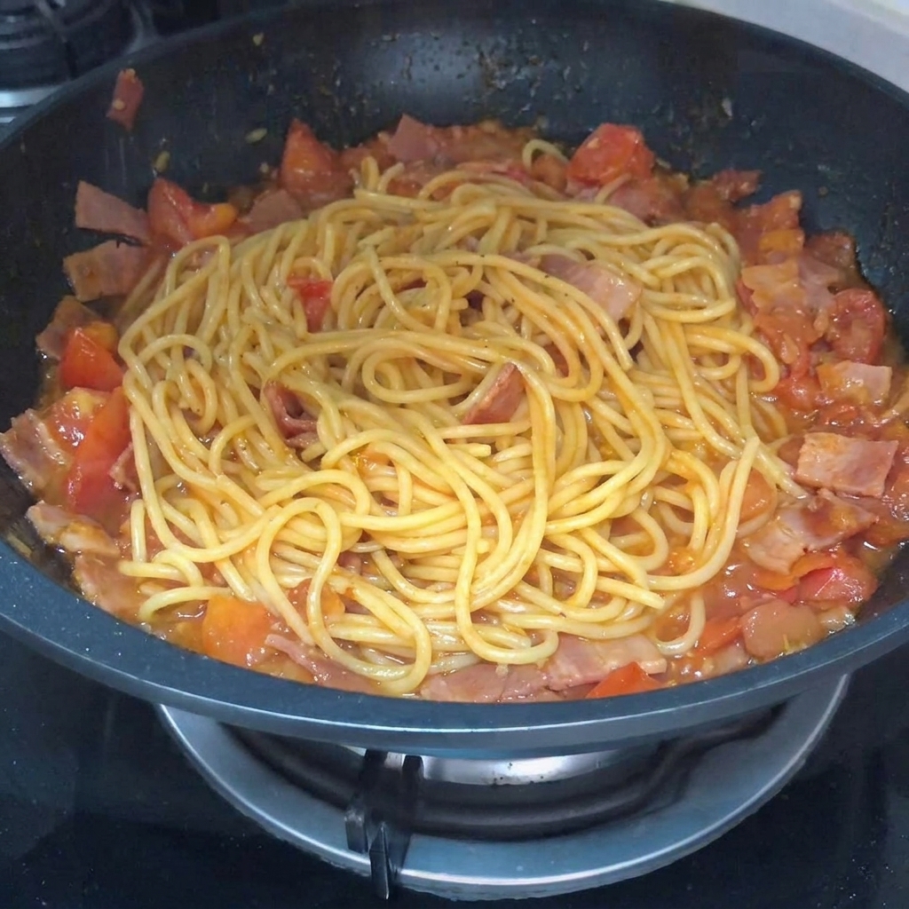A close-up of pasta being tossed in a pan with tomato and bacon sauce, with black pepper being sprinkled over the top.
