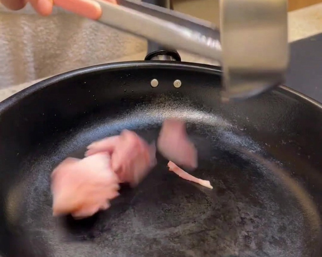 Adding bacon pieces into a black skillet to fry.