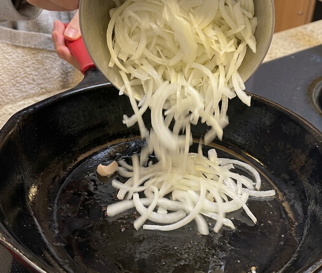 Pouring thinly sliced onions from a pot into a black skillet.