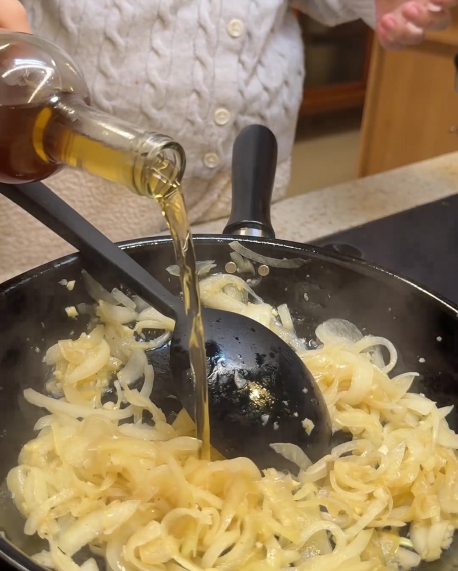White wine and balsamic vinegar being poured into a pan filled with sautéed onions and garlic.
