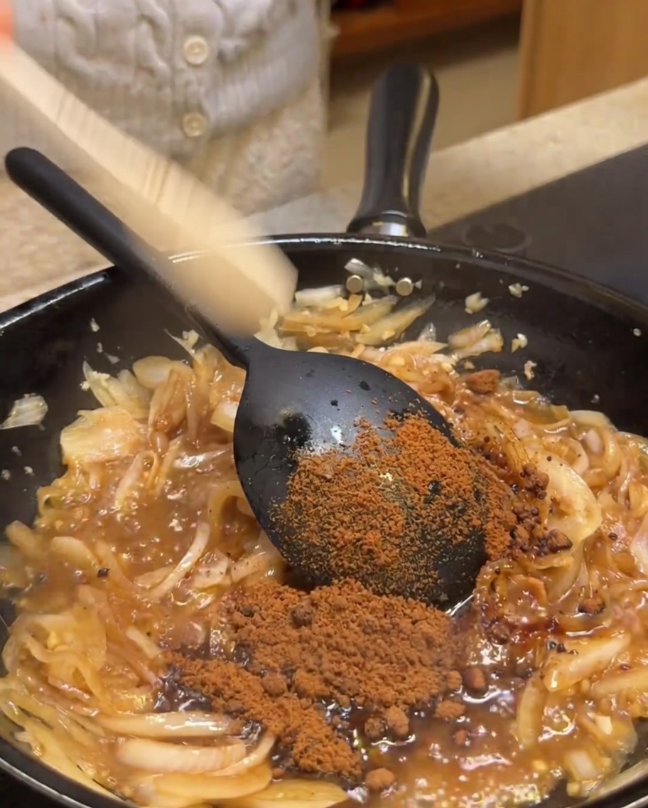Brown sugar being sprinkled into a skillet containing sautéed onions and aromatics.