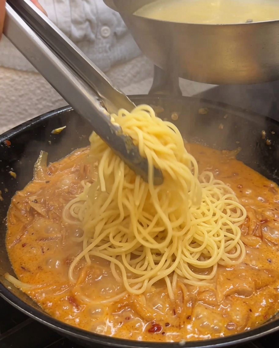 Cooked spaghetti being tossed in a creamy onion sauce using metal tongs.