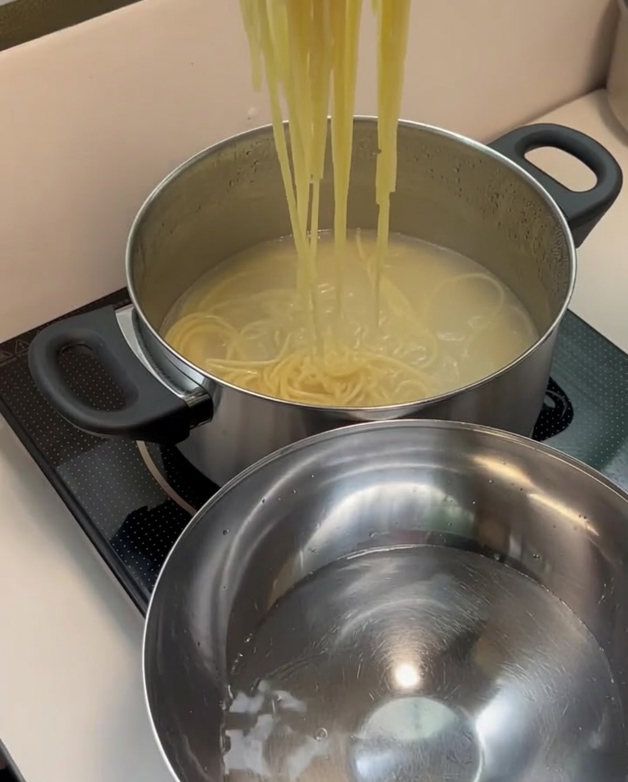Strained cooked spaghetti noodles being moved into a stainless steel bowl containing fresh, clear cold water.
