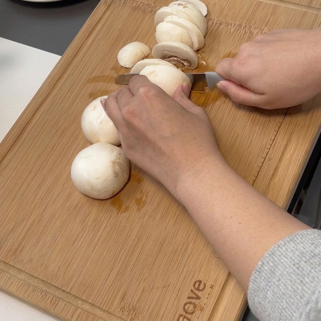 A hand using a chefs knife to thinly slice white button mushrooms on a bamboo cutting board.