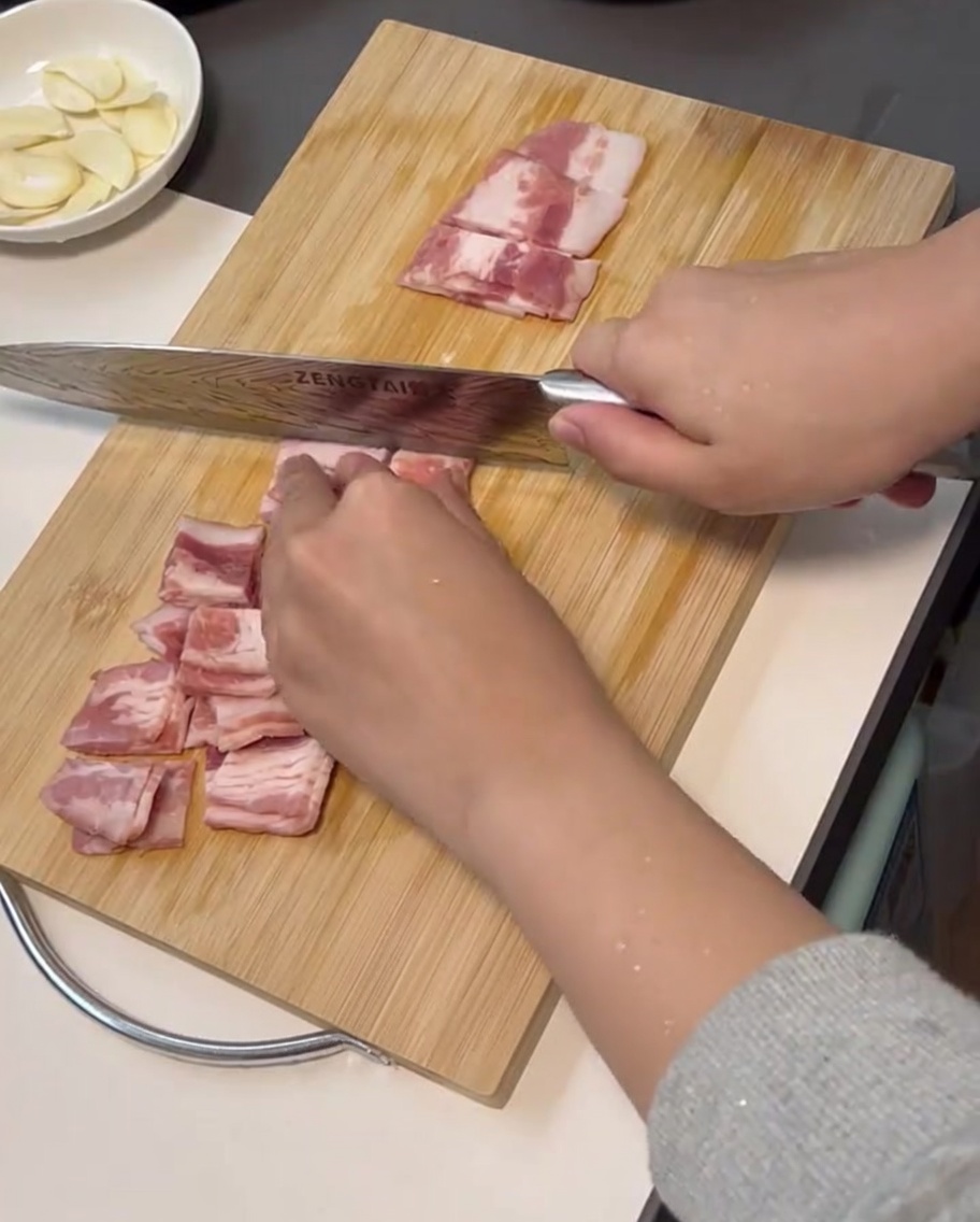 Fresh bacon strips being cut into small, even pieces on a cutting board using a sharp knife.