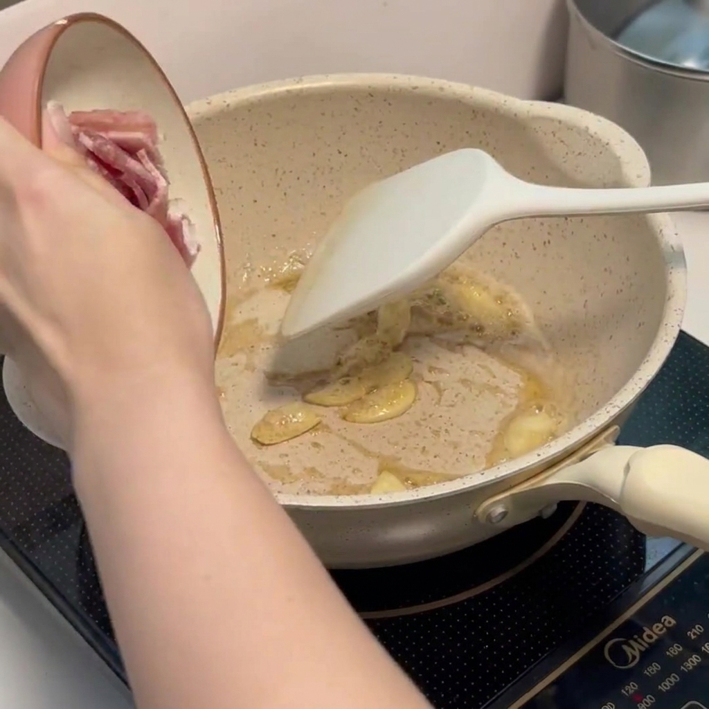 Small pieces of chopped bacon frying in a pan alongside fragrant garlic slices.