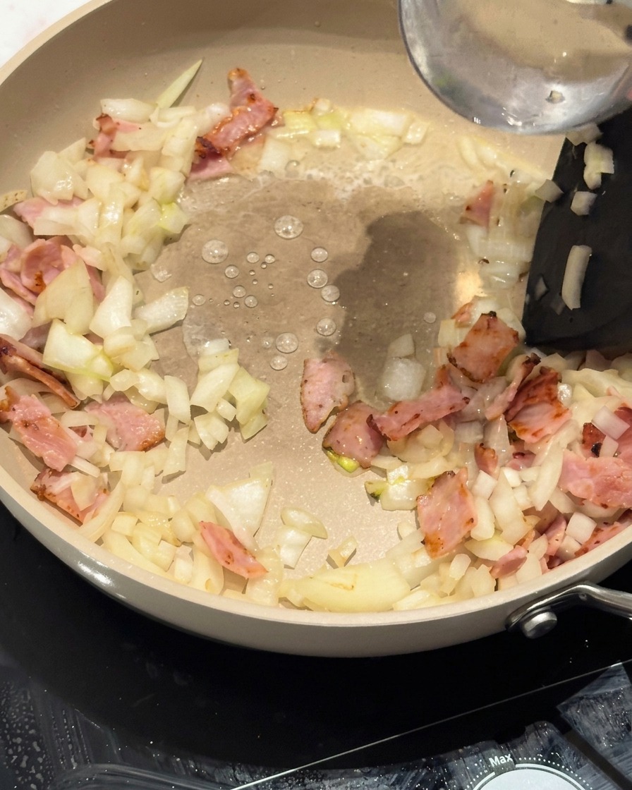 Curly noodles being placed into the pan over the bacon and onion base.