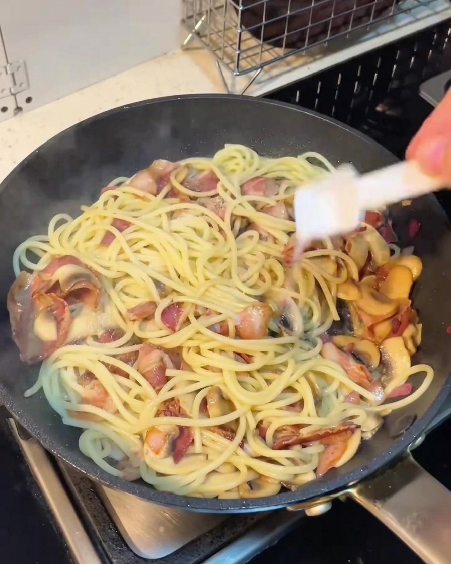 A hand holding a shaker over a pan of spaghetti with bacon and mushrooms, seasoning the dish.