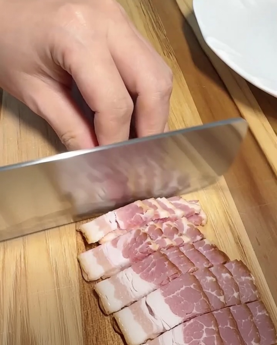 A close-up of bacon strips being sliced into small, even pieces on a wooden surface.
