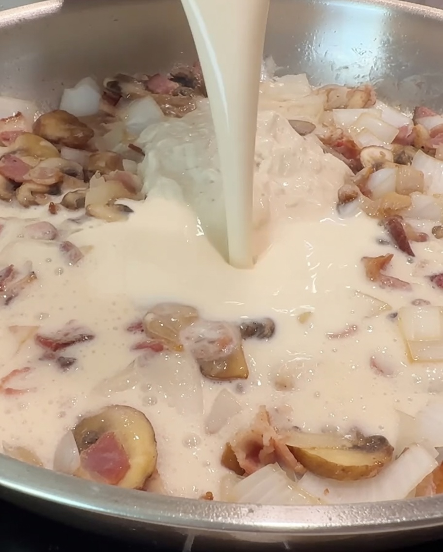 A cup of milk being poured into the pan to blend with the Alfredo sauce and sautéed ingredients.