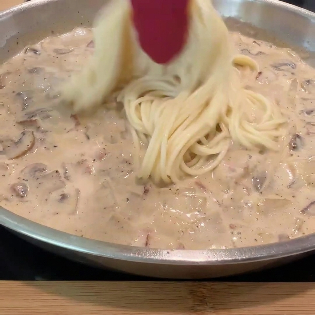 Cooked spaghetti noodles being added to the pan of creamy sauce and ingredients.