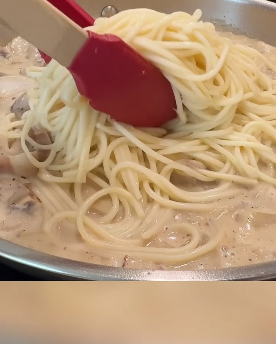 Spaghetti being tossed with creamy sauce, bacon, and mushrooms in a pan using red tongs.