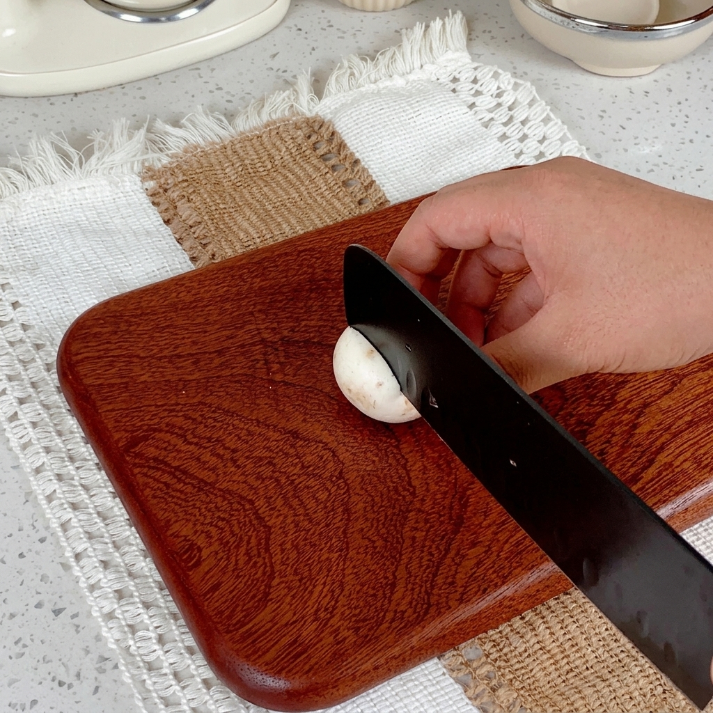 A hand using a black knife to slice a white button mushroom on a wooden cutting board.