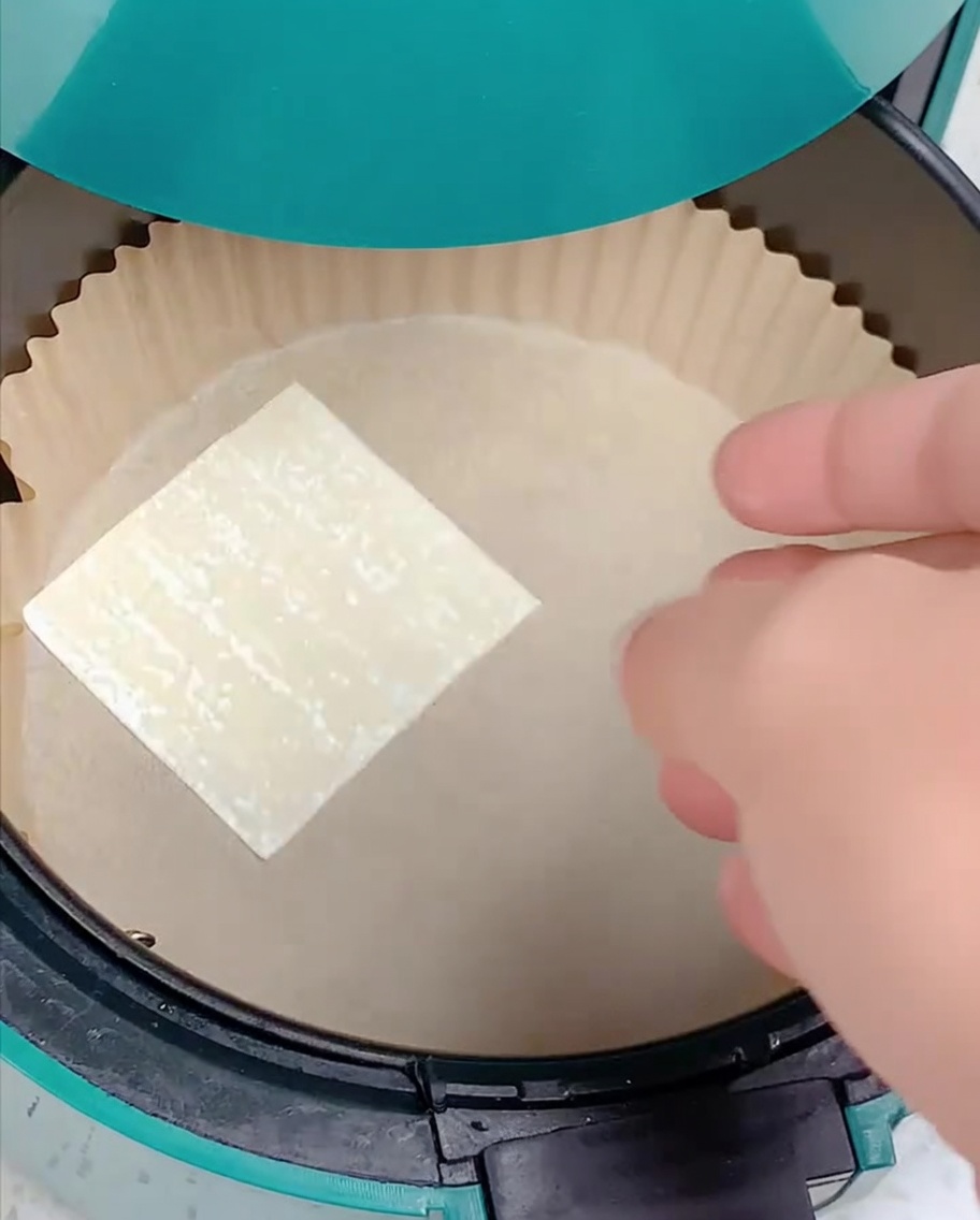 A hand placing a square wonton wrapper onto a parchment-lined air fryer basket.