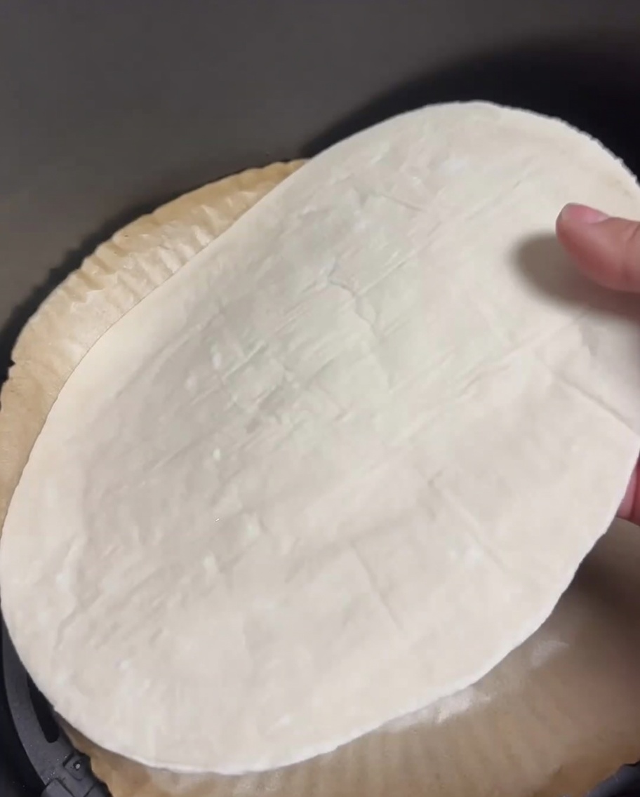 A raw scallion pancake laying flat in an air fryer basket lined with parchment paper.
