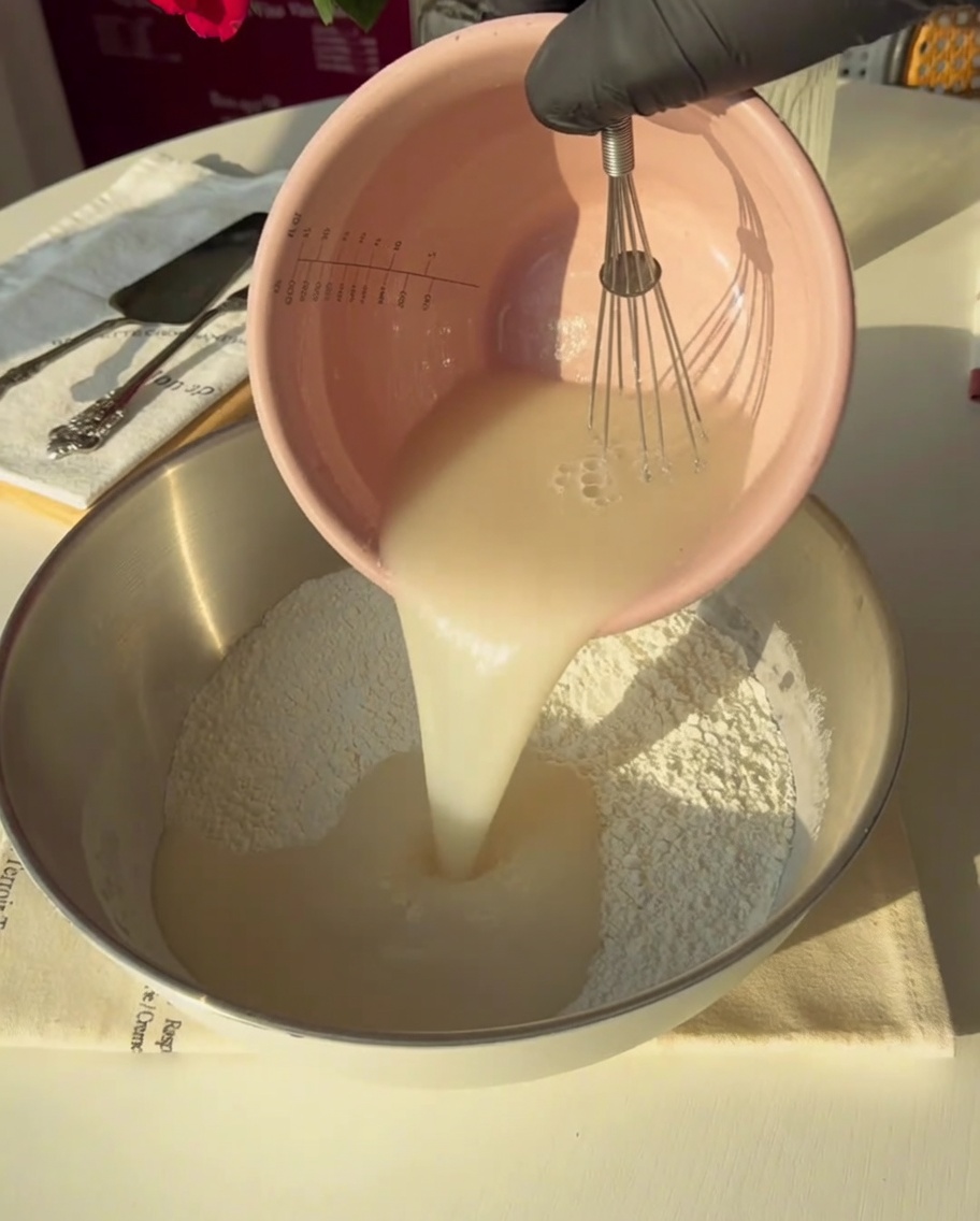 A gloved hand pouring the yeast-water mixture into a large metal bowl containing flour.