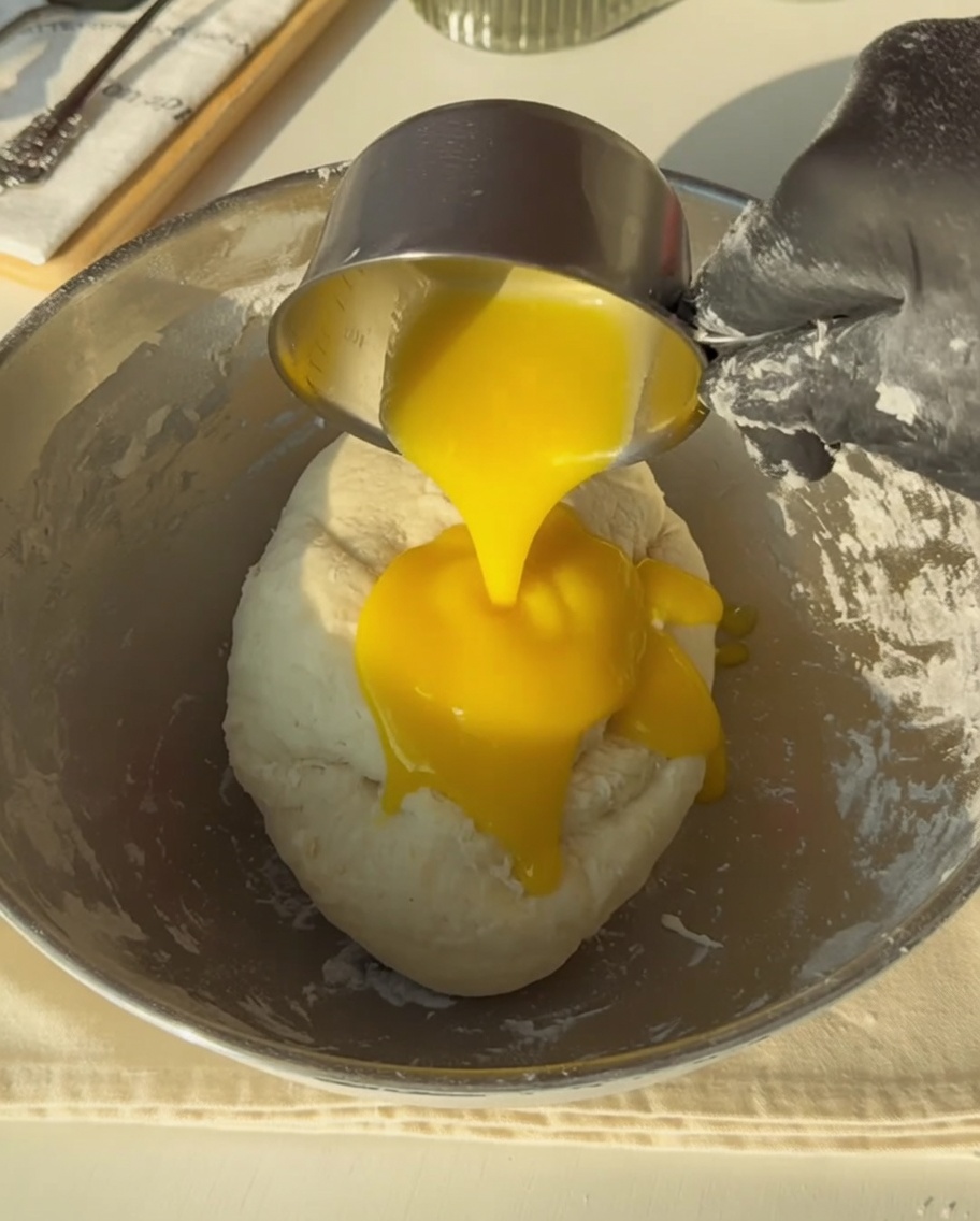 Gloved hand pouring golden melted butter from a measuring cup onto the dough.