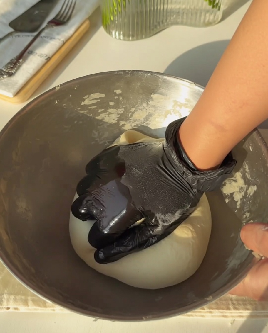 A gloved hand firmly kneading the dough in a metal bowl until it is smooth and uniform.