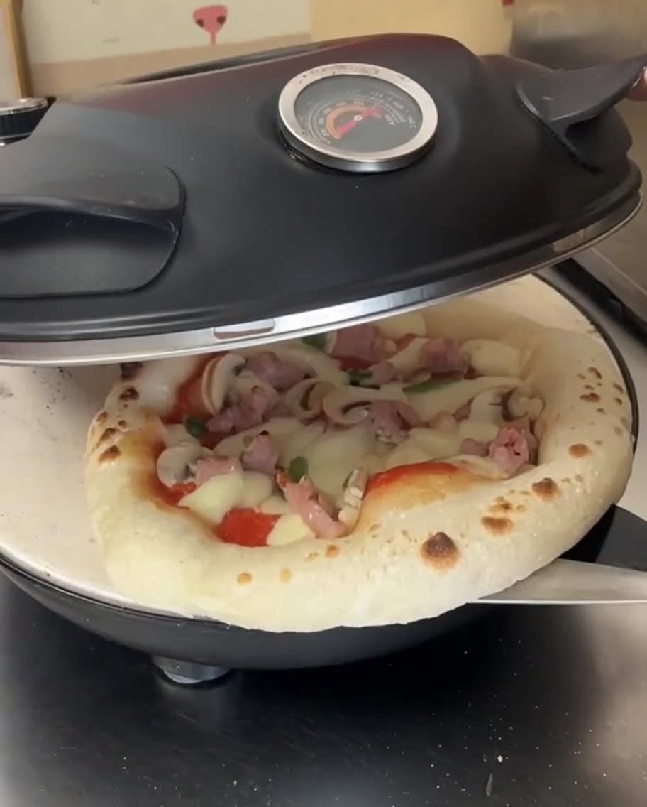 A partially baked pizza with melted cheese inside an open pizza oven, being rotated by a metal pizza peel.