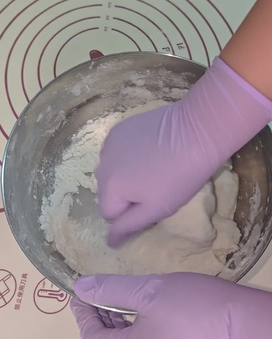 Hands wearing purple gloves kneading the dough in a metal bowl.