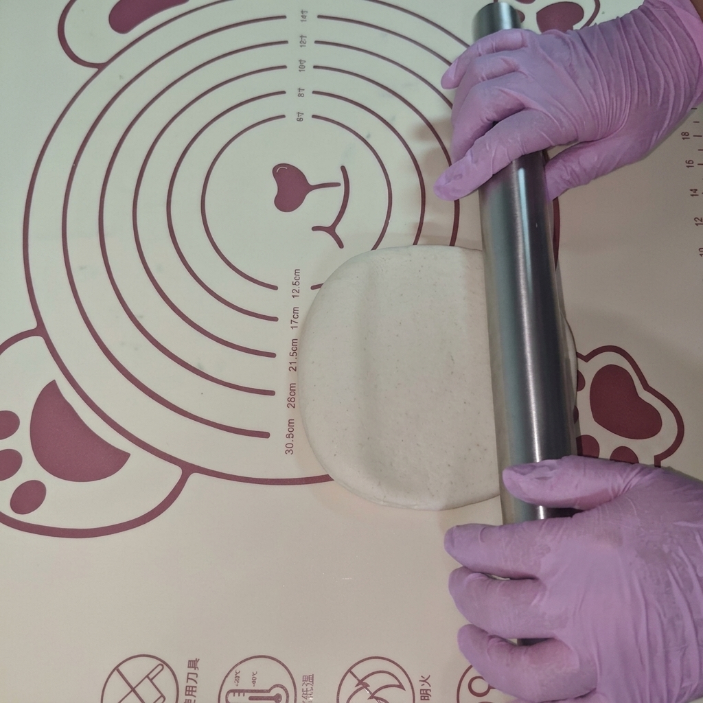 A piece of dough being rolled into a flat circle on a silicone mat using a rolling pin.