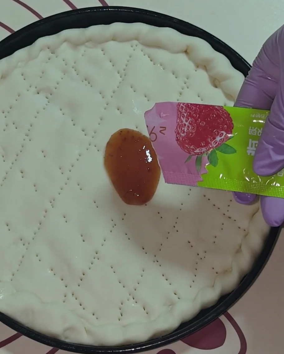 A close-up view of a hand in a purple glove spreading red strawberry jam onto the circular pizza dough using a small packet.