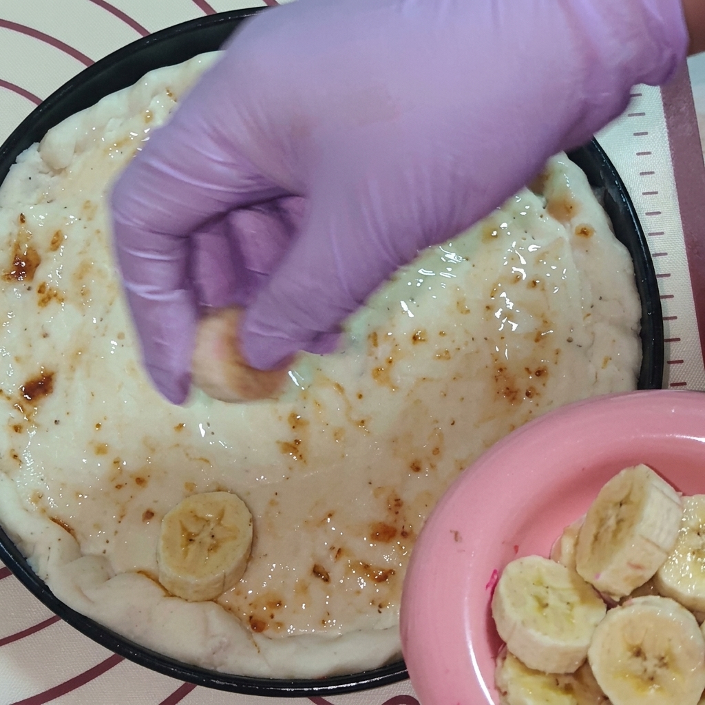 A hand wearing a purple glove placing slices of fresh fruit, including banana and dragon fruit, onto the jam-covered pizza dough.