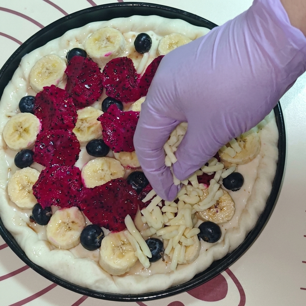 A top-down view of a hand wearing a purple glove sprinkling shredded mozzarella cheese over a fruit-topped pizza.