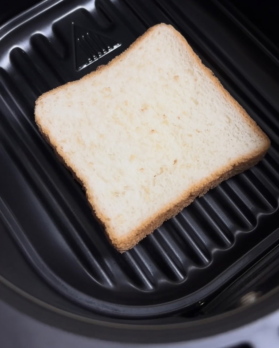 A plain slice of white bread sitting flat in the bottom of an air fryer basket.