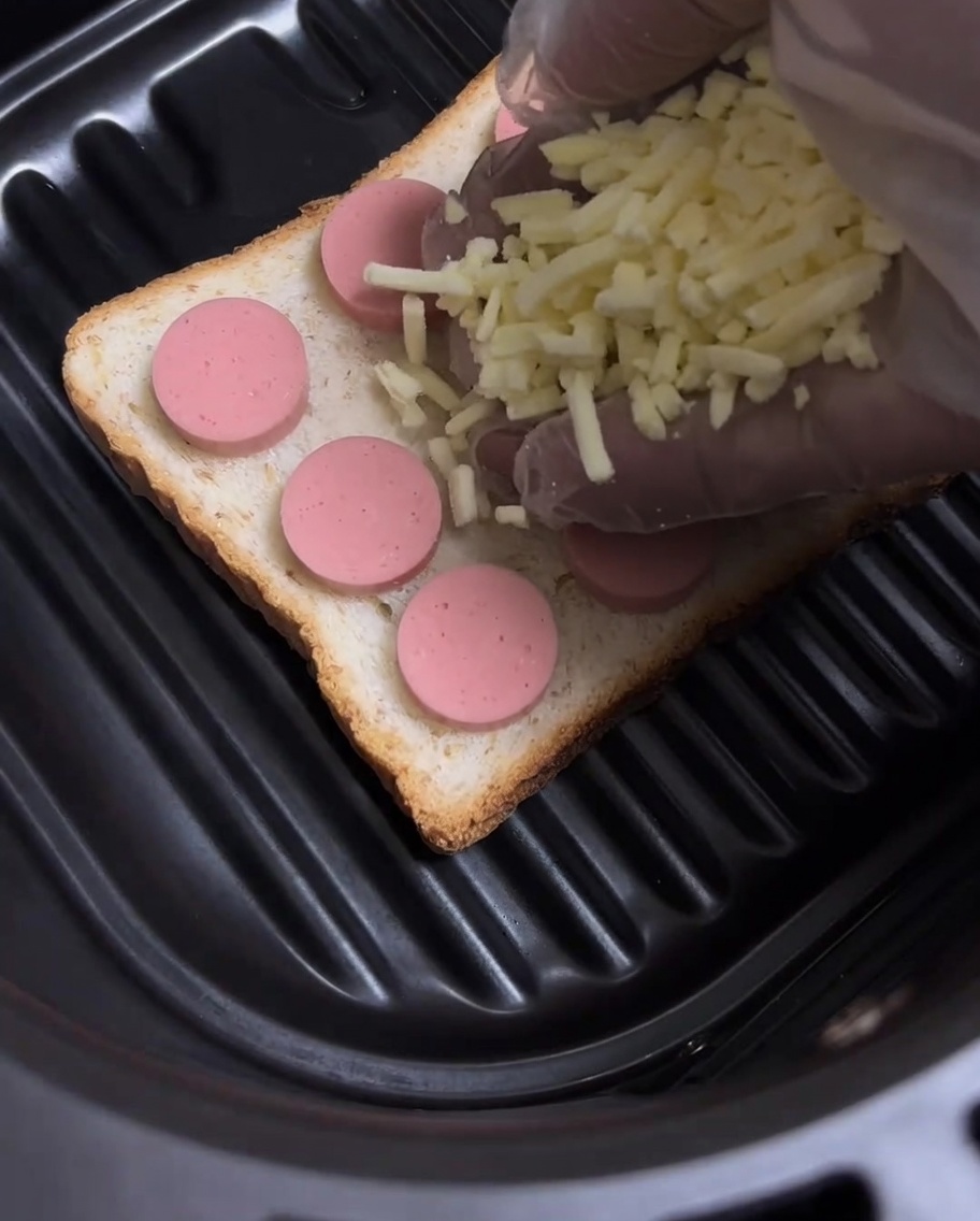 Shredded yellow cheese being sprinkled over the hot dog slices on the toast.
