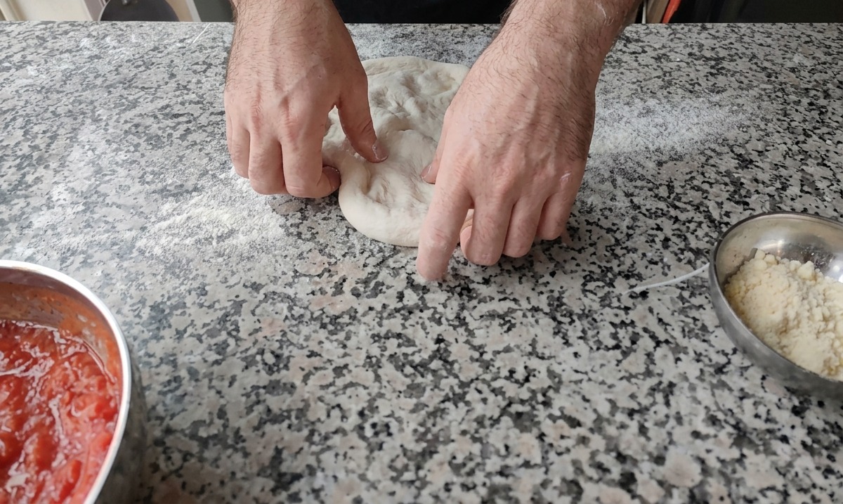 Two hands pressing down on the center of a round pizza dough ball on a lightly floured marble surface.