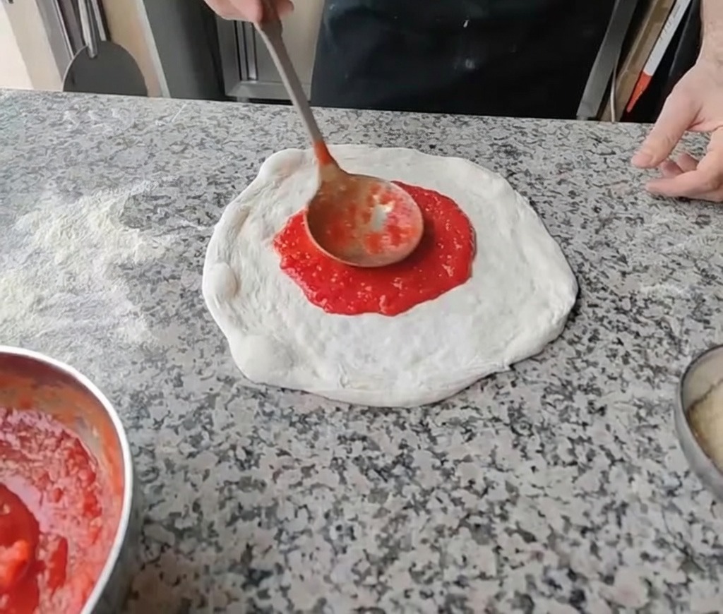 Spreading bright red tomato sauce in the center of the stretched pizza dough with a ladle.