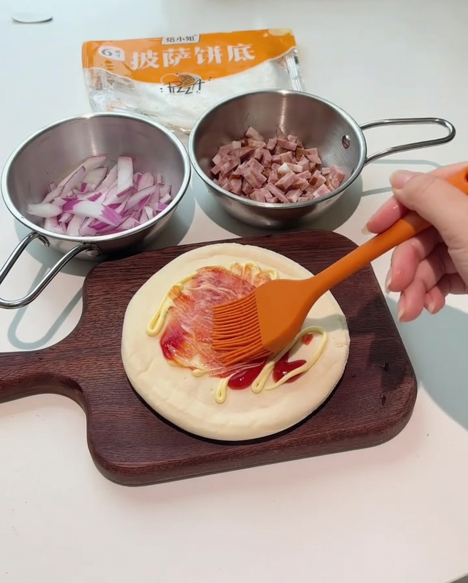 An orange silicone brush spreading red and white sauces over the pizza base.