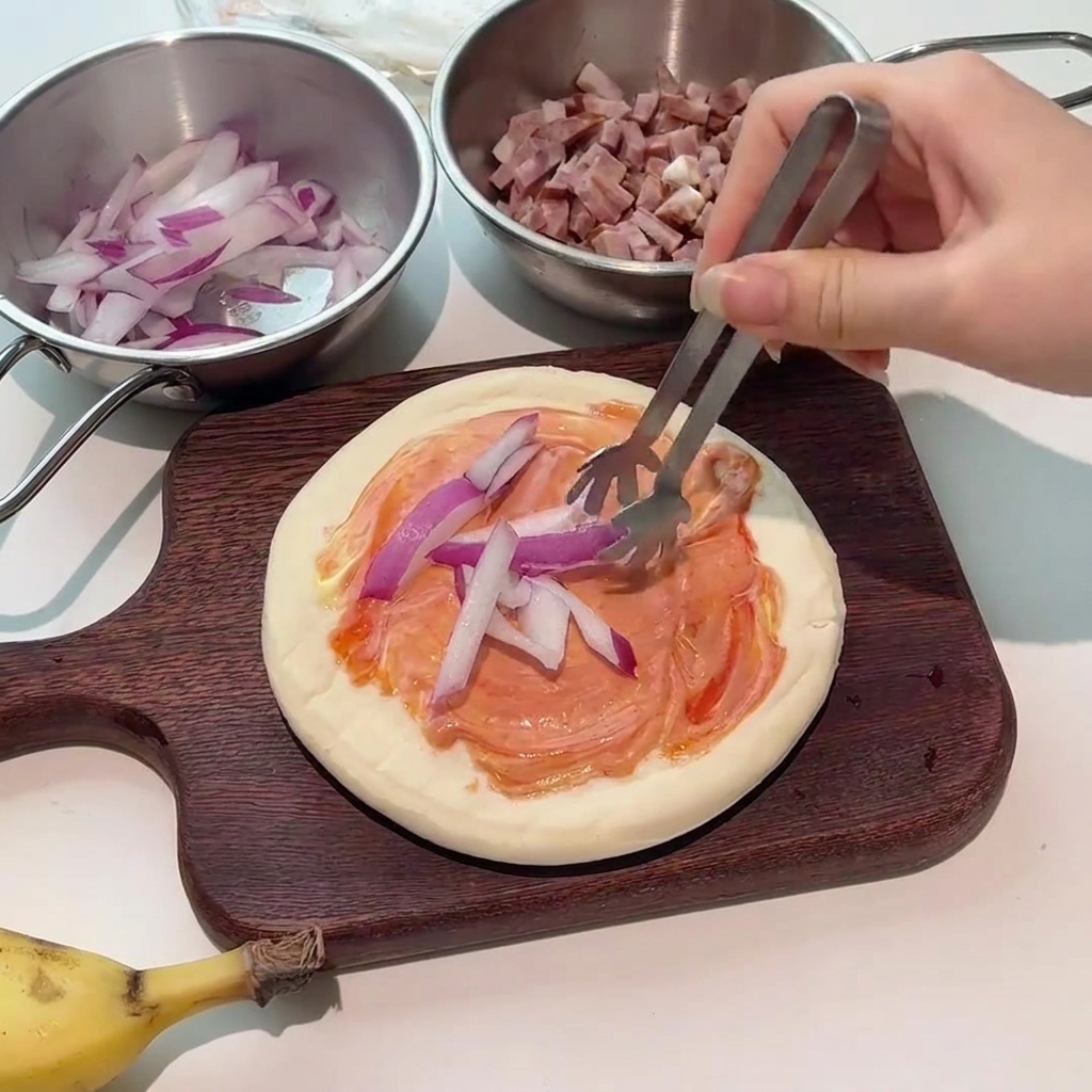 Tongs placing fresh red onion slices onto the sauce-coated pizza crust.