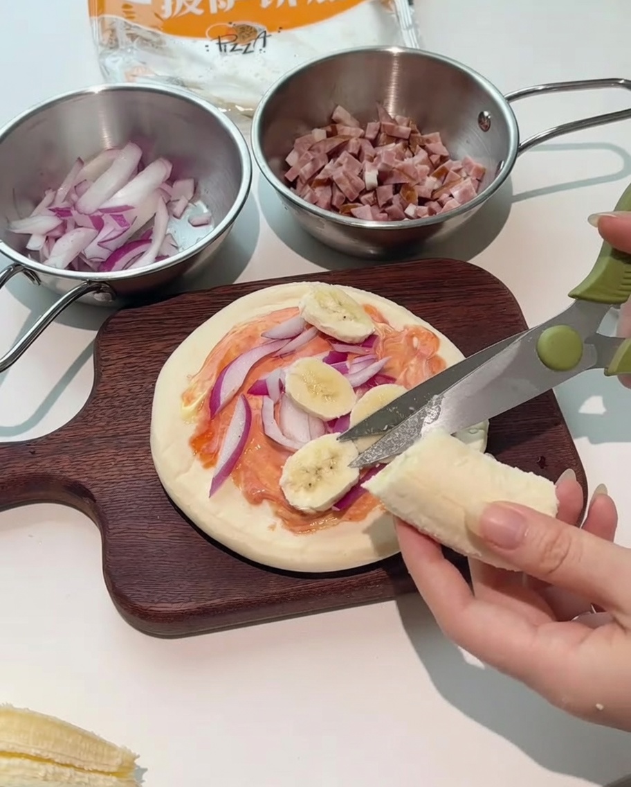 Kitchen scissors cutting fresh banana slices directly onto the pizza assembly.