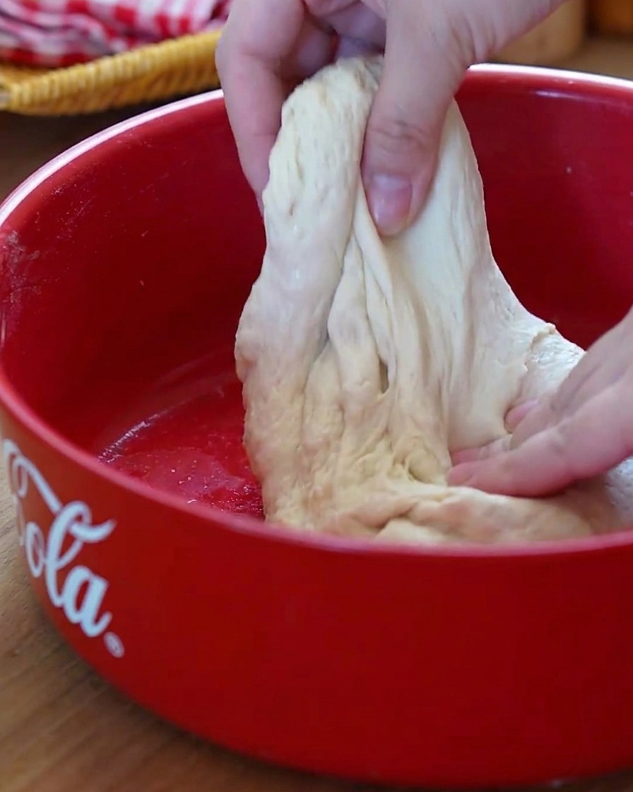 A close-up view of hands performing the stretch-and-fold technique on soft pizza dough inside a red bowl.