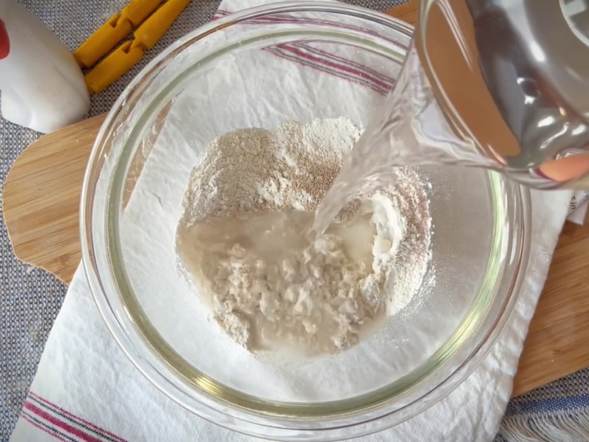 A glass bowl containing a mixture of flour, yeast, salt, and water being combined with a spatula.
