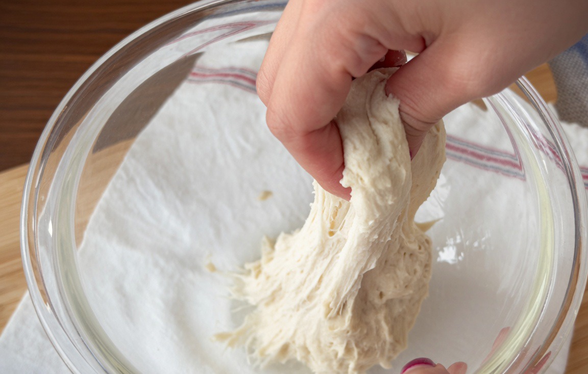 A pair of hands with wet skin stretching and folding high-hydration dough within a glass bowl.