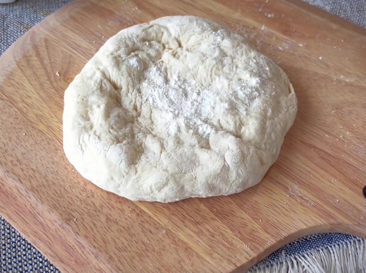 A round piece of pizza dough being stretched on a wooden surface, with a clearly defined thick outer rim.