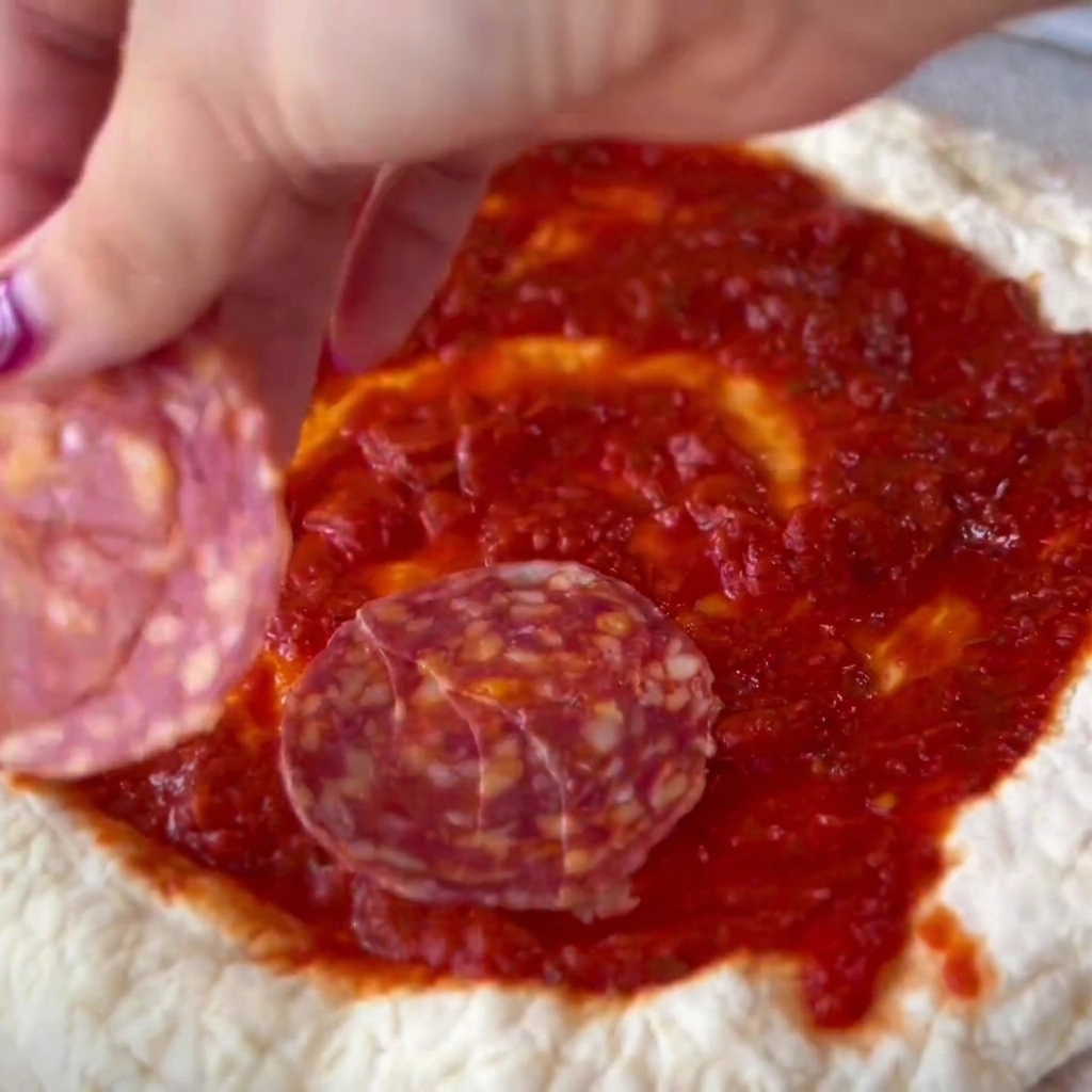 Slices of salami being carefully placed onto the sauced center of a pizza dough.