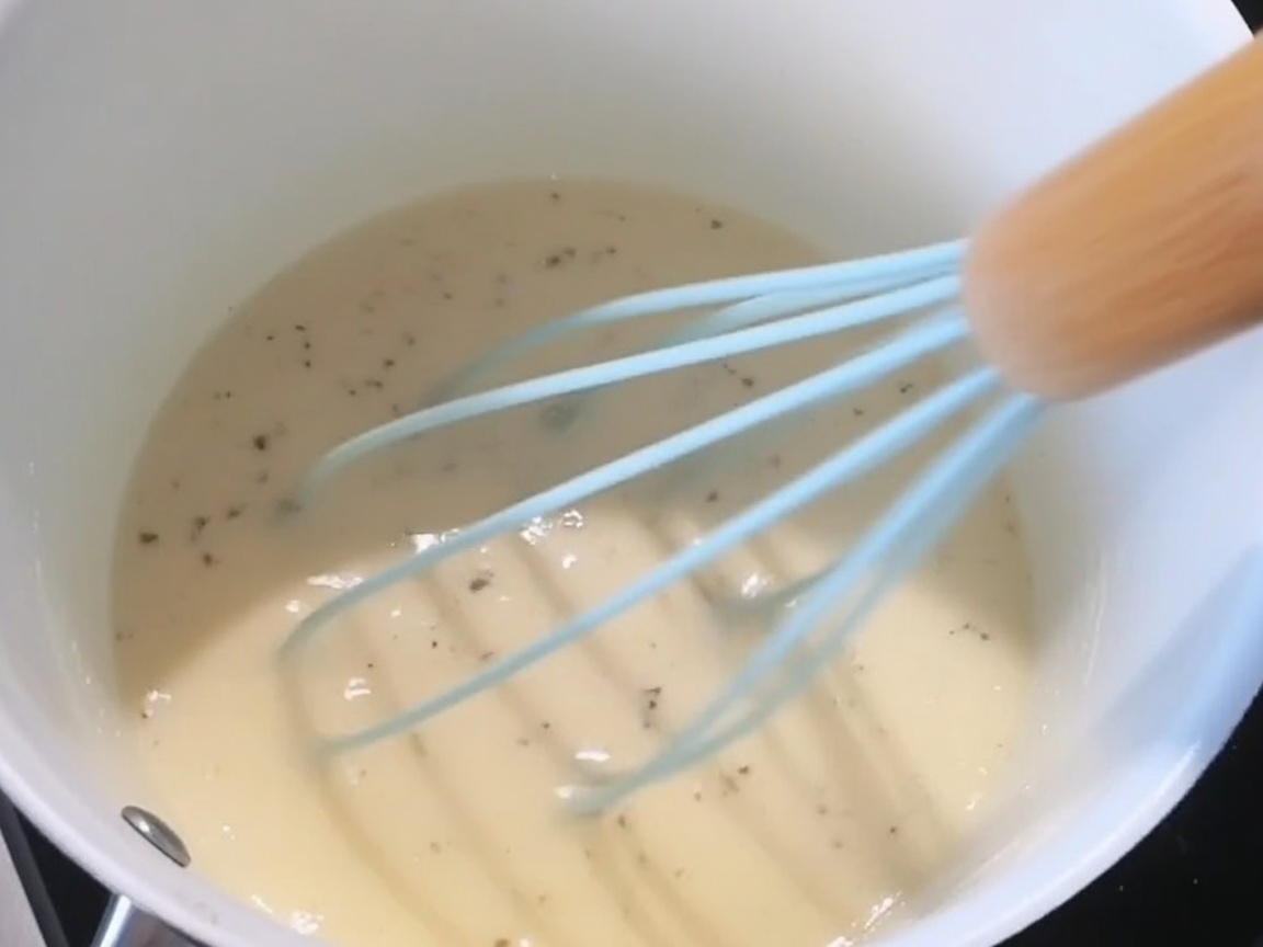Salt, black pepper, and shredded cheese being added to the creamy white sauce.