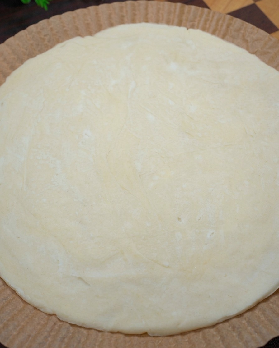 A close-up of hands shaping the edges of a raw, circular scallion pancake on parchment paper to form a pizza crust.