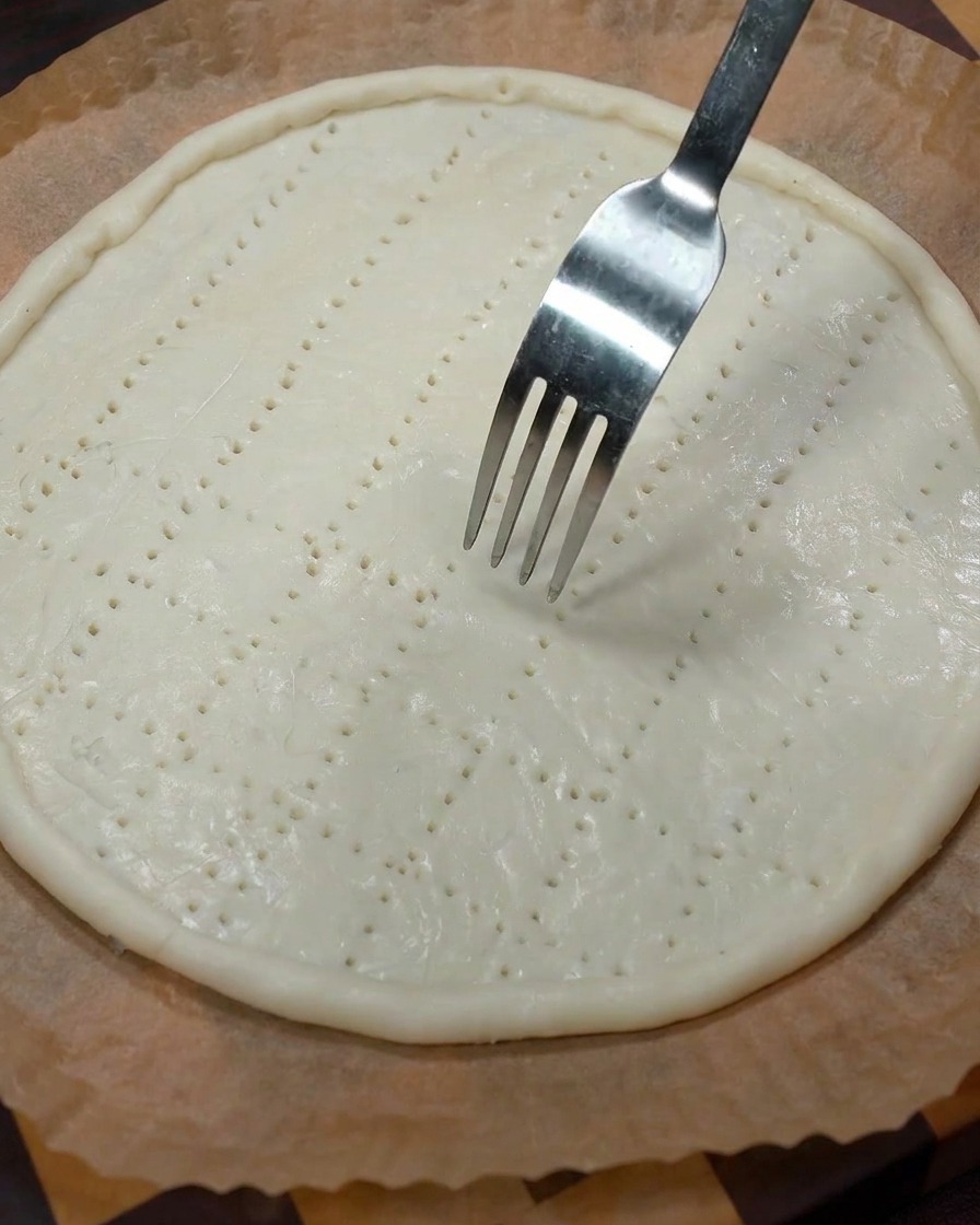 A metal fork pressing into the raw dough base to create a grid of ventilation holes.